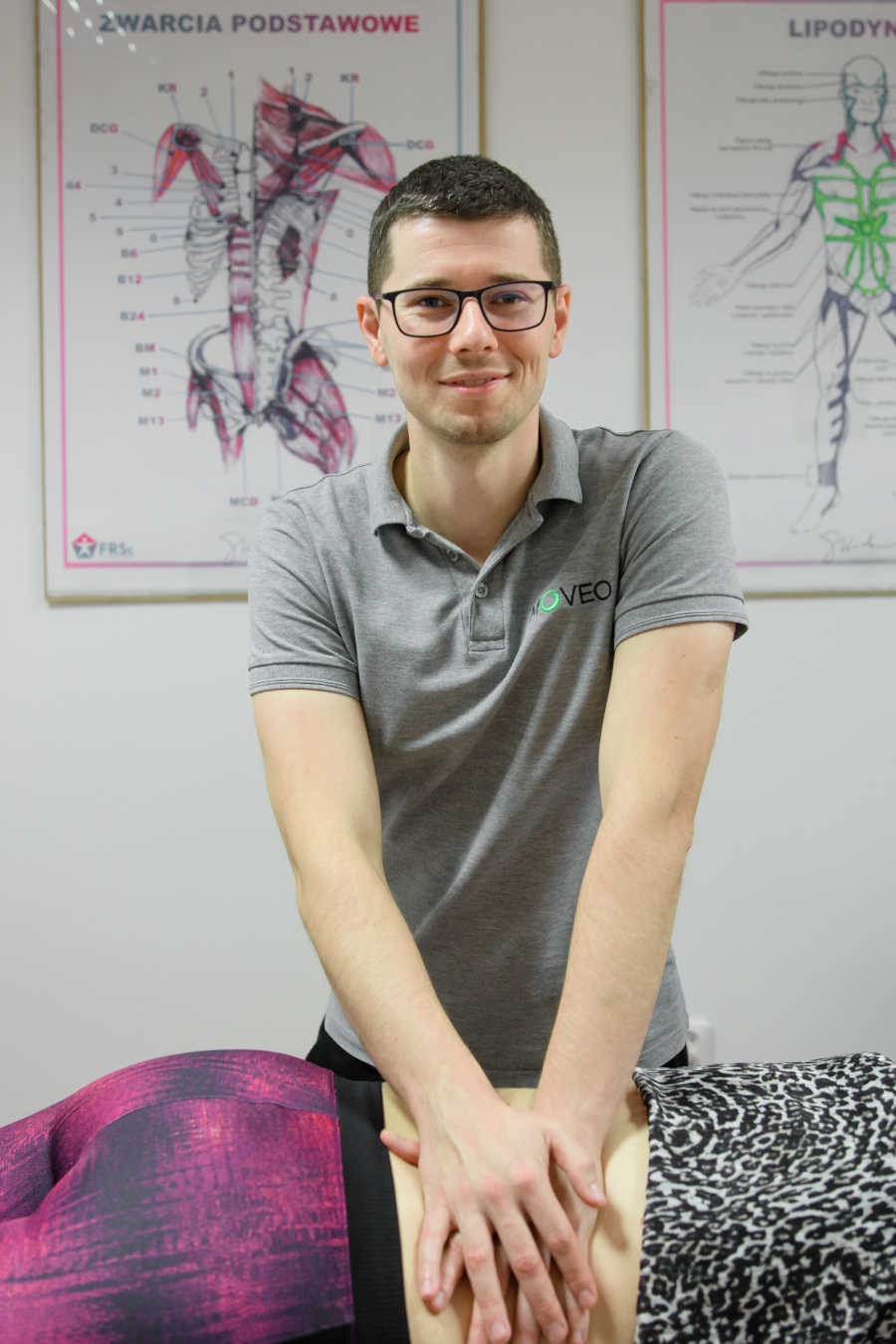 Terapeuta w szarej koszulce polo z logo masuje plecy pacjentki leżącej na stole; w tle plakaty anatomiczne.