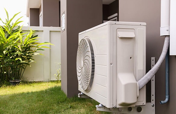 Zewnętrzna jednostka klimatyzacji zamontowana na ścianie budynku, widoczna na tle trawnika i roślinności.