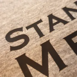 Fragment beżowej koszulki z czarnym nadrukiem dużych liter widziany pod kątem, ukazujący strukturę materiału.