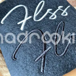 Ciemnoszary kawałek materiału z wyhaftowanymi białymi i czarnymi napisami, leżący na sznurku jutowym.