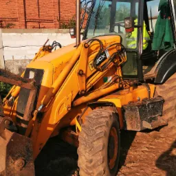 Żółta koparko-ładowarka JCB 3CX z brudnymi oponami i łyżką, zaparkowana na ziemi, w tle ceglany budynek z krzyżem na szczycie
