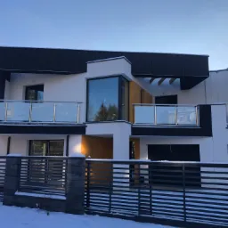 Nowoczesny, dwukondygnacyjny dom z płaskim dachem i białymi ścianami, wykończony czarnymi panelami elewacyjnymi, widoczny zza szarego ogrodzenia w zimowej scenerii.