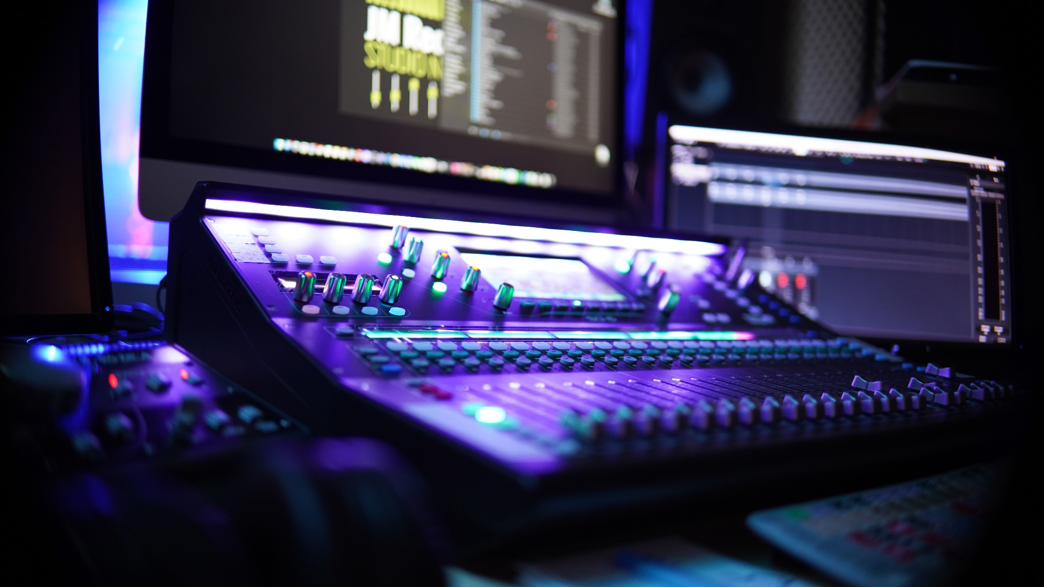 Profesjonalny mikser audio z podświetleniem LED w odcieniach fioletu i zieleni, widoczne monitory z oprogramowaniem do edycji dźwięku w tle.