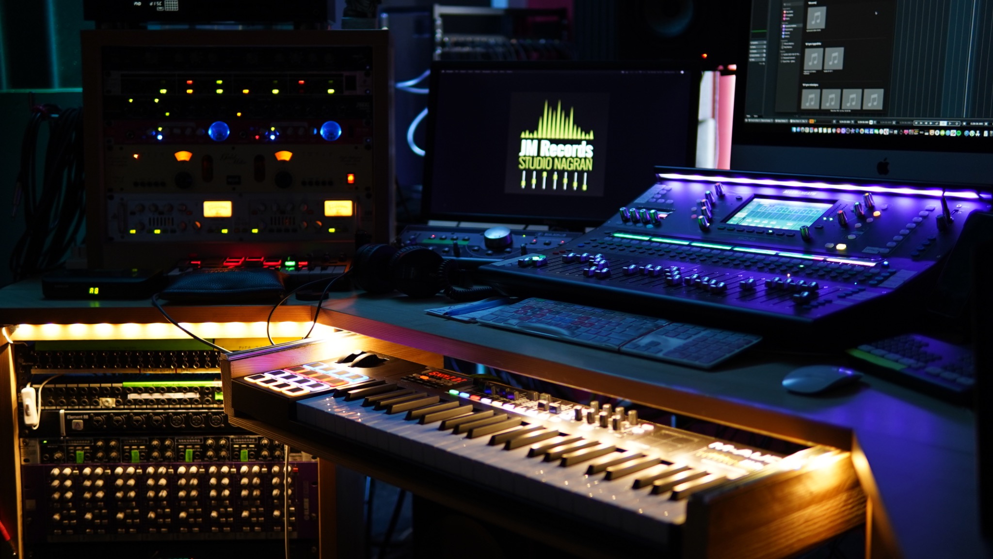Profesjonalny mikser audio z podświetleniem LED, syntezator klawiszowy, monitory studyjne z logo na ekranie w ciemnym pomieszczeniu.