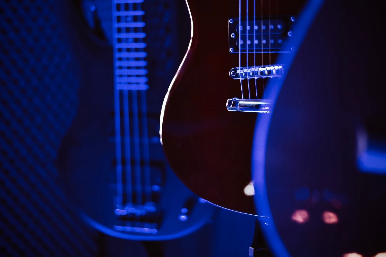 Trzy gitary elektryczne w niebieskim oświetleniu, z widocznymi strunami i elementami korpusu, ustawione w pomieszczeniu z wytłumieniem akustycznym.