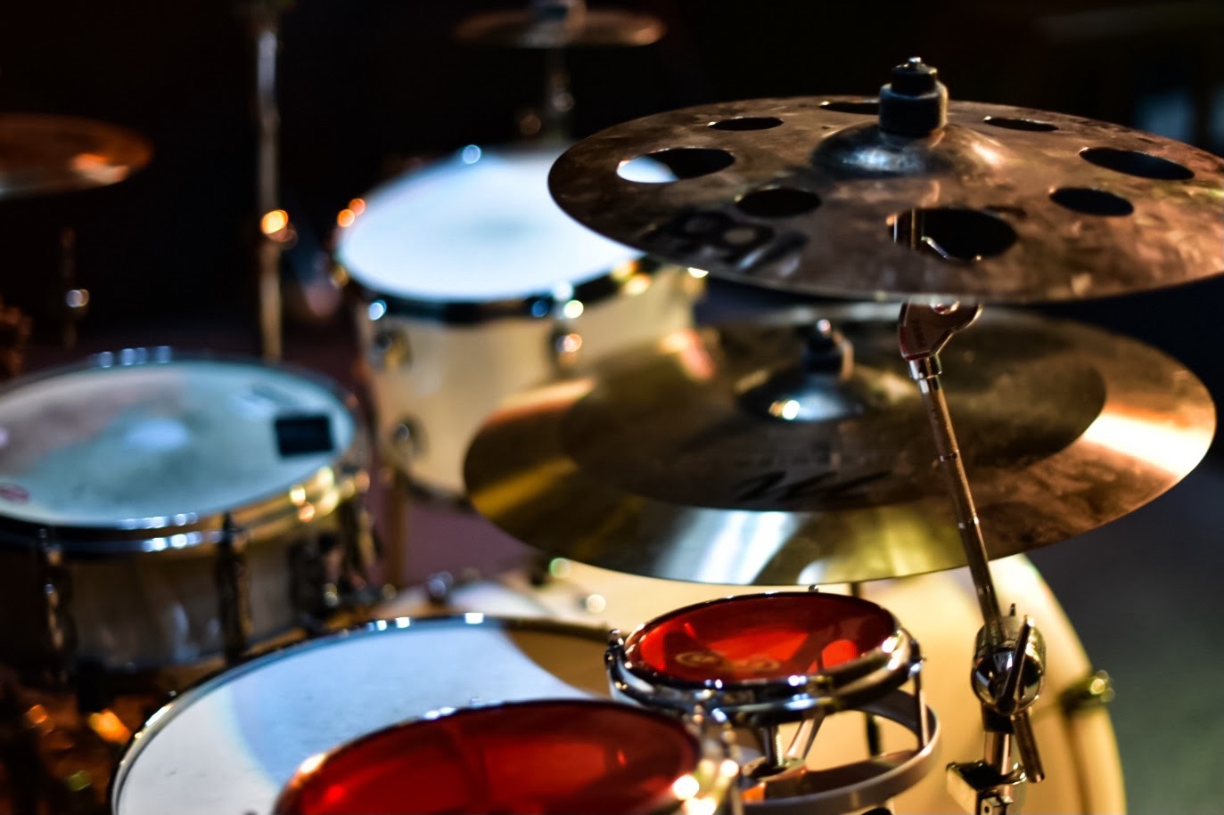 Zestaw perkusyjny z talerzami oświetlony punktowym światłem, widoczne bębny o białych i czerwonych membranach.