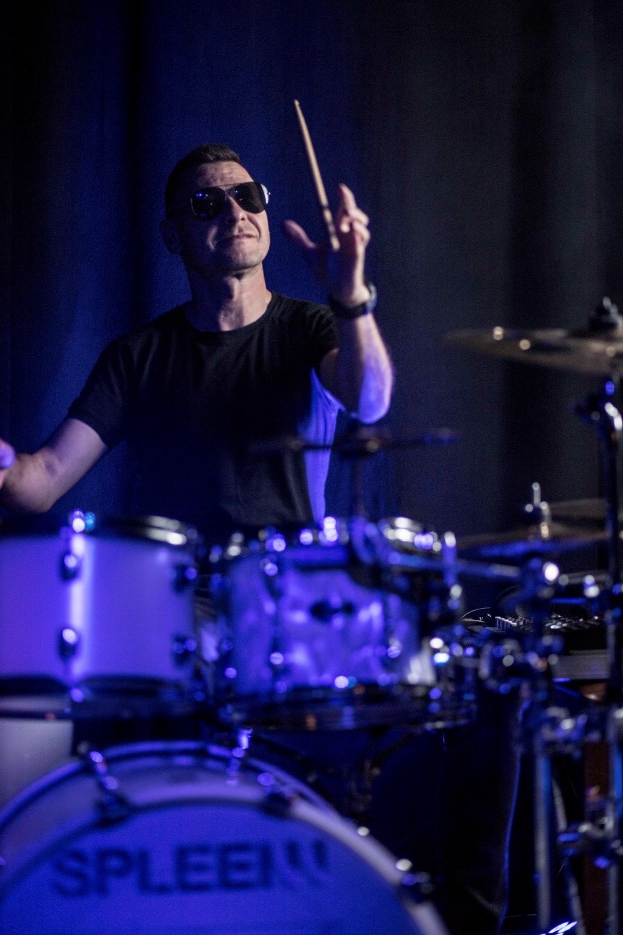 Perkusista w okularach przeciwsłonecznych, z podniesioną pałeczką, grający na perkusji pod niebieskim oświetleniem scenicznym, widoczny fragment zestawu perkusyjnego marki 'Spleen'.