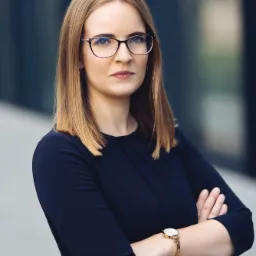 Portret profesjonalnej kobiety w okularach, z założonymi rękami, ubrana w granatową sukienkę i złoty zegarek na tle nowoczesnego budynku.