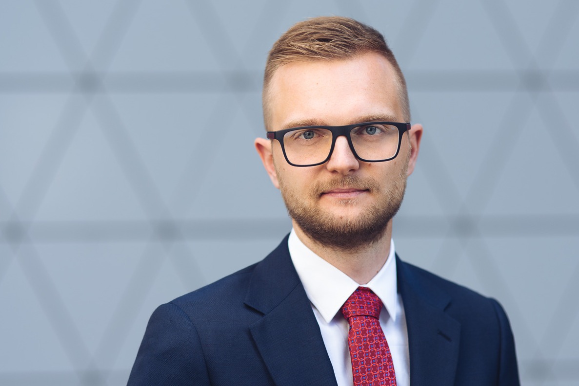 Portret mężczyzny w garniturze i okularach na tle szarej ściany z geometrycznym wzorem.