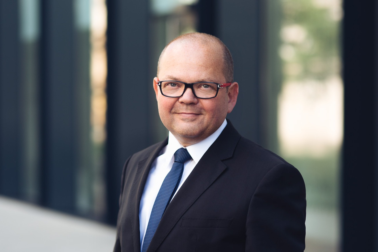 Portret uśmiechniętego mężczyzny w okularach i garniturze z niebieskim krawatem na tle nowoczesnego budynku.