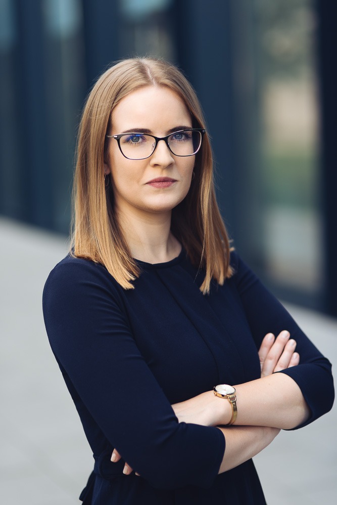 Portret profesjonalnej kobiety w okularach, z założonymi rękami, ubrana w granatową sukienkę i złoty zegarek na tle nowoczesnego budynku.