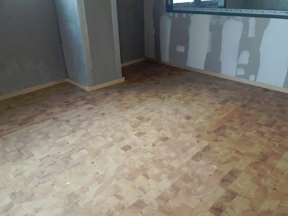 Ułożona podłoga z drewnianych klocków w surowym stanie, widoczne niedokończone ściany z oknem w tle.