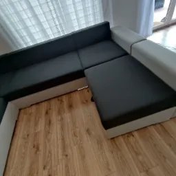 Częściowo rozłożona narożna kanapa w odcieniach szarości i bieli na drewnianej podłodze, widoczna z góry, z zasłoniętym oknem w tle.