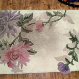 Jasny dywan z motywem kwiatowym w odcieniach różu, fioletu i zieleni, leżący na drewnianej podłodze, z fragmentem czarnego węża odkurzacza widocznym w tle.