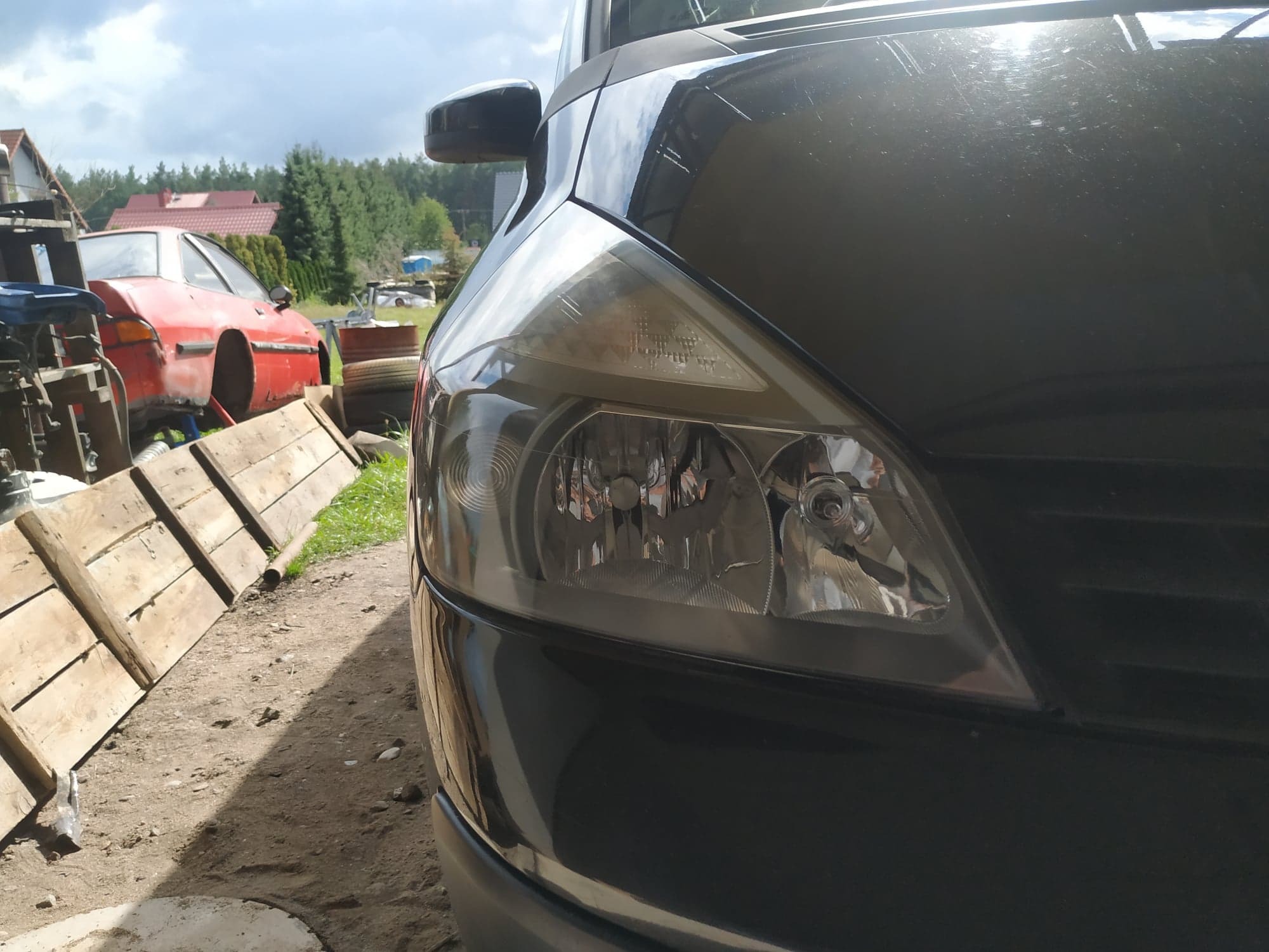 Reflektor i fragment karoserii czarnego samochodu na tle warsztatu z czerwonym autem w trakcie renowacji.