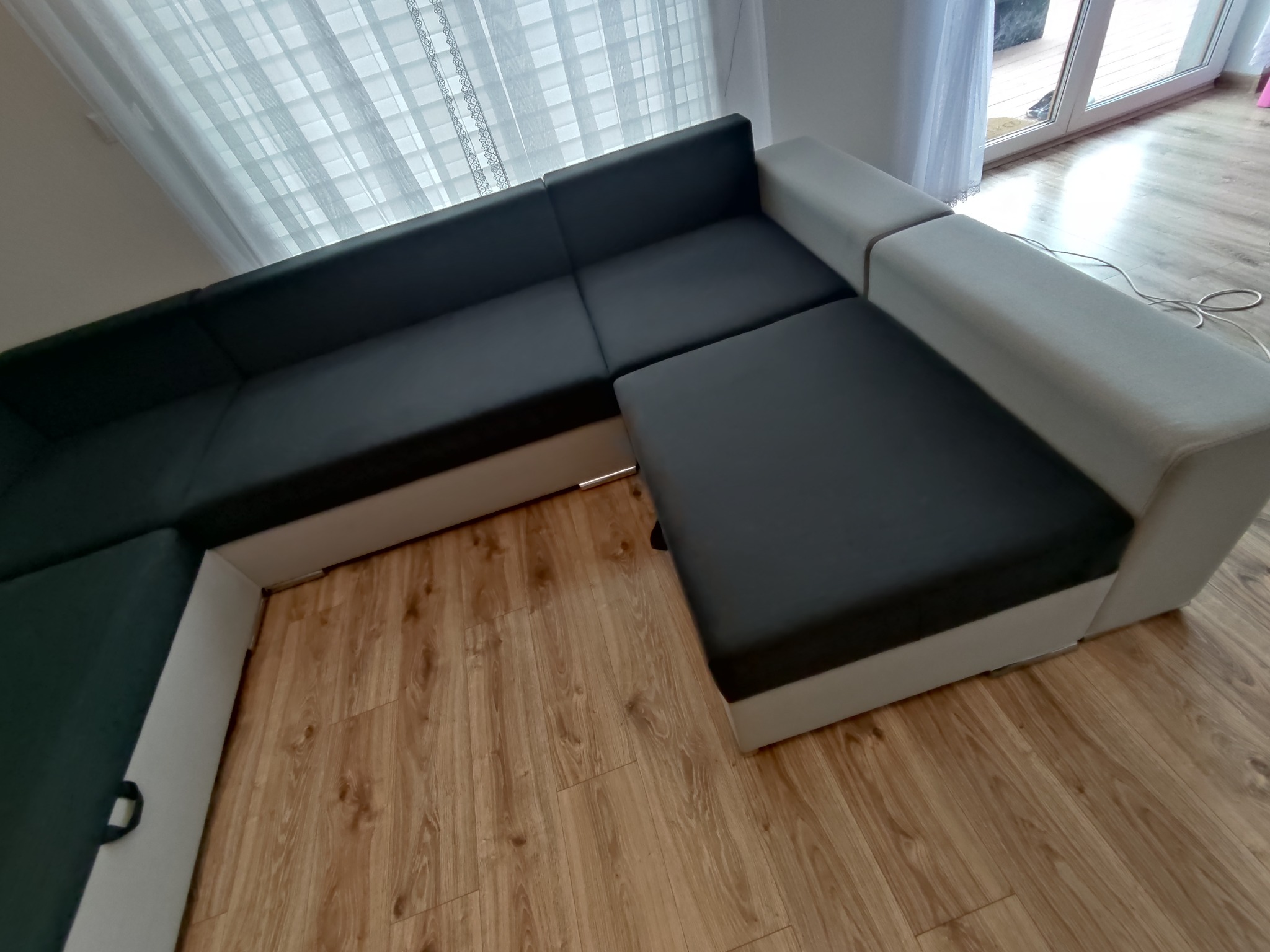 Częściowo rozłożona narożna kanapa w odcieniach szarości i bieli na drewnianej podłodze, widoczna z góry, z zasłoniętym oknem w tle.