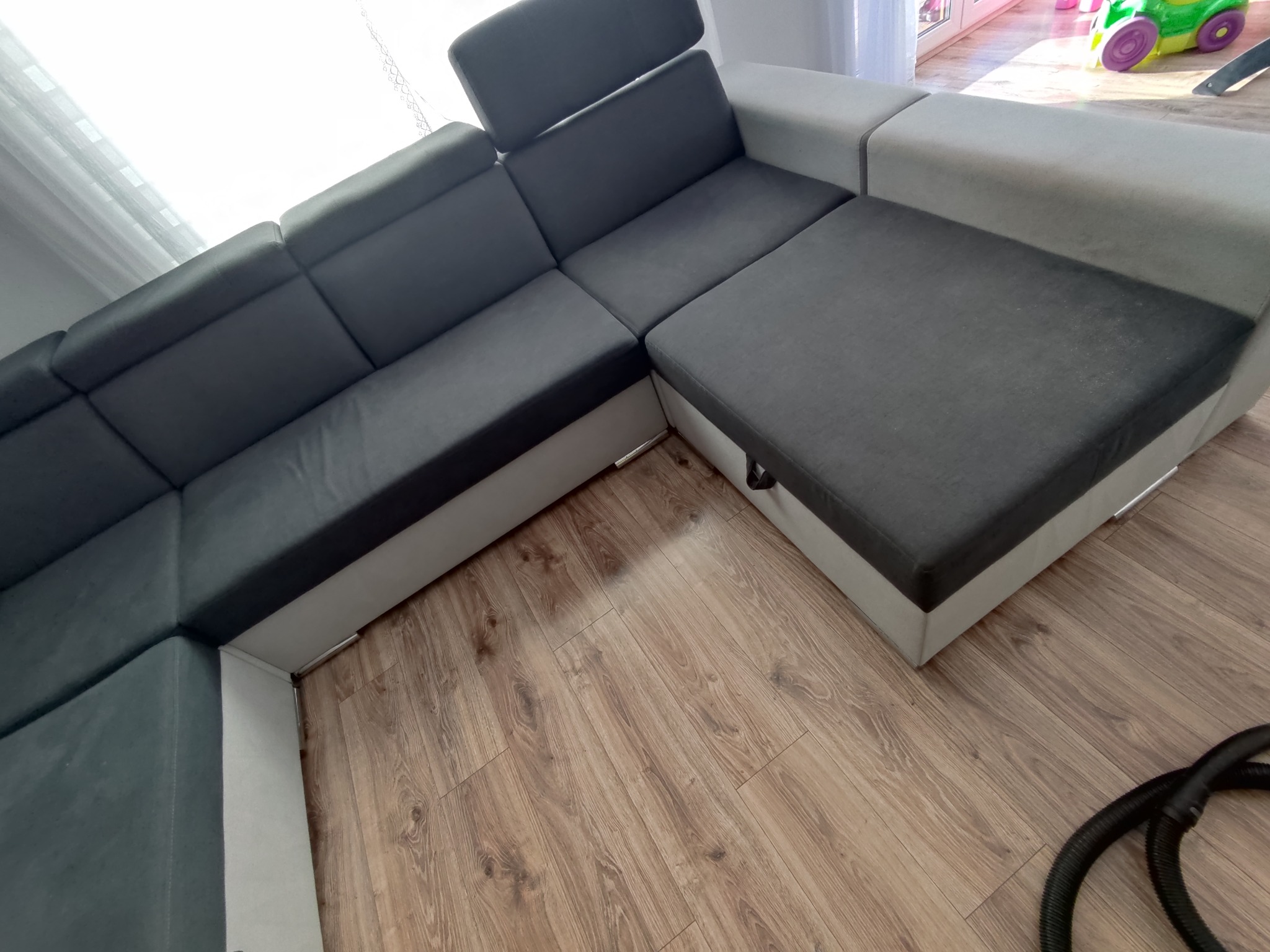 Szary narożnik z funkcją spania i pojemnikiem na pościel, widoczny fragment węża odkurzacza na podłodze.