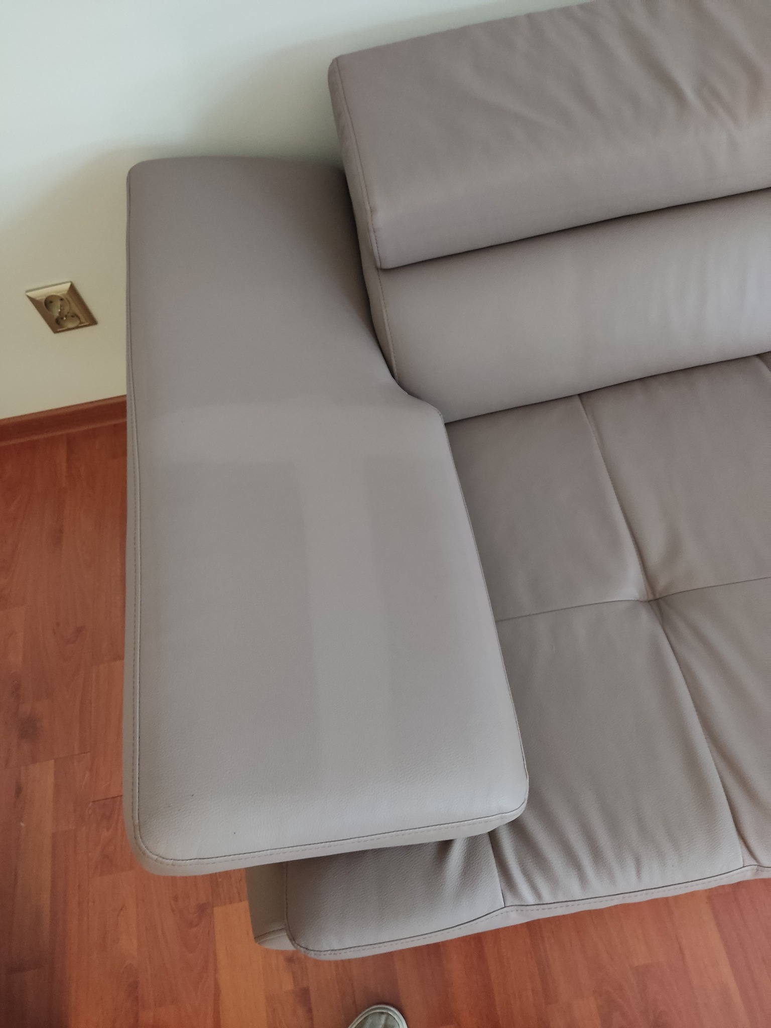 Fragment beżowej skórzanej kanapy z widocznym podłokietnikiem i pikowanym siedziskiem, sfotografowany z góry w naturalnym świetle, z gniazdkiem elektrycznym widocznym na ścianie w tle.