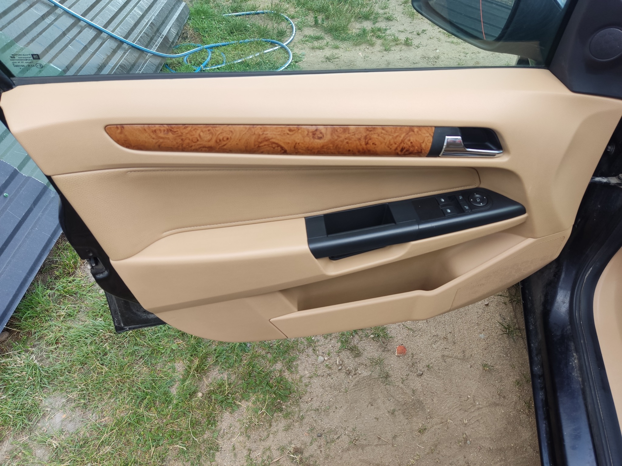 Wnętrze drzwi samochodu z beżową tapicerką i drewnopodobnym wykończeniem, widoczne elementy sterowania szybami i lusterkami, tło z blachy falistej i trawy.