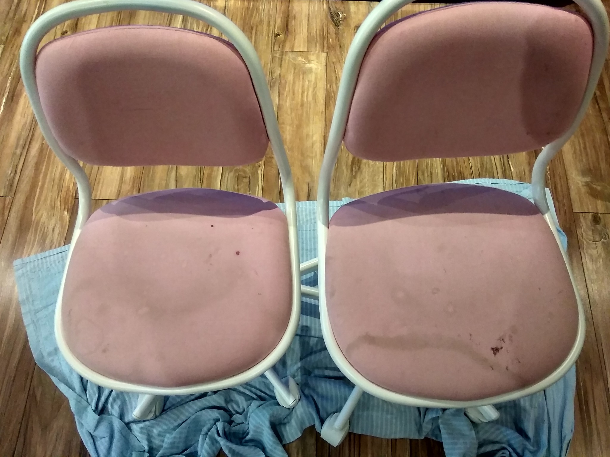 Dwa tapicerowane krzesła w odcieniach różu, z widocznymi plamami na siedziskach, ustawione na niebieskiej, pogniecionej tkaninie na tle drewnianej podłogi.