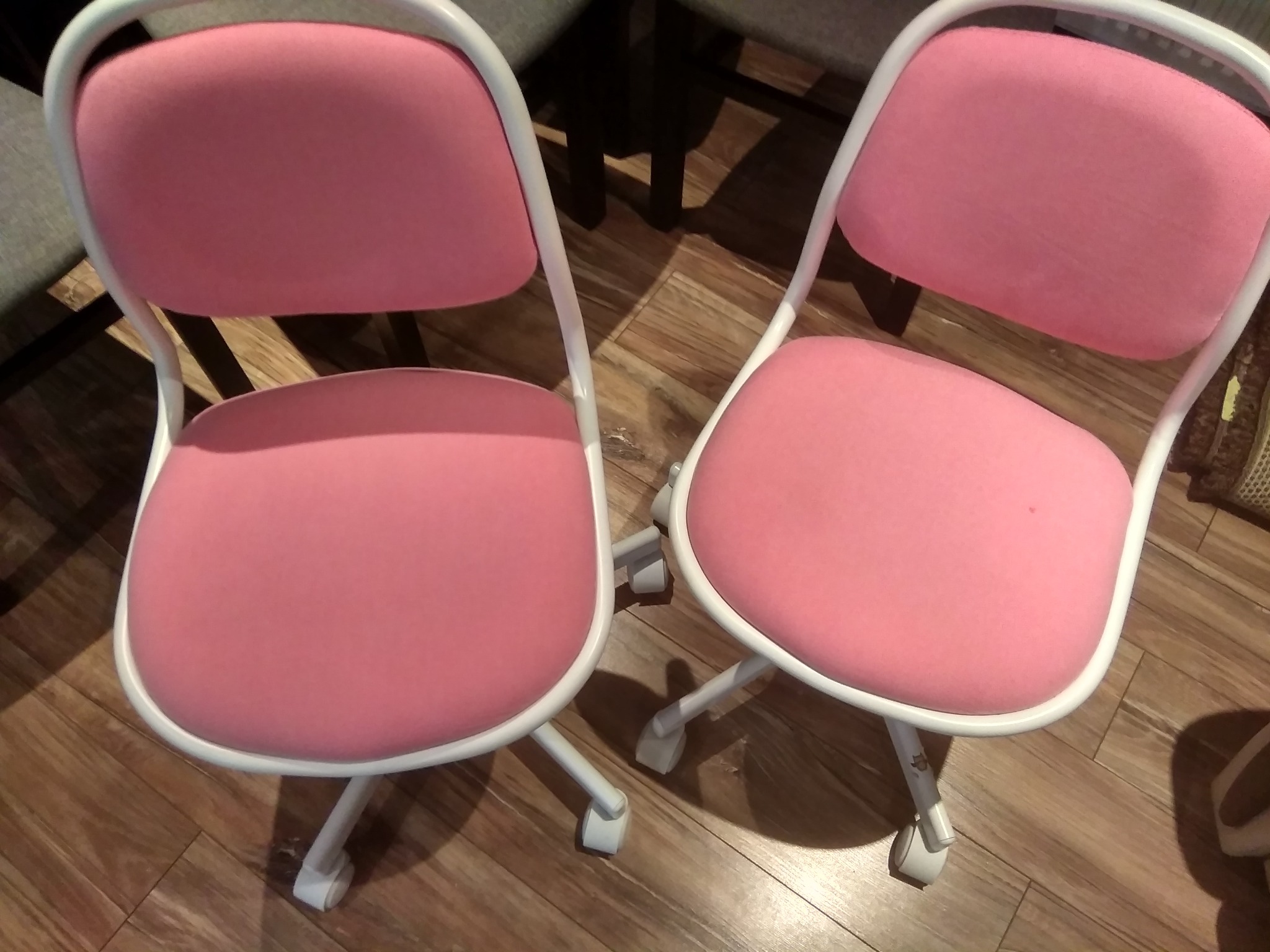 Dwa różowe krzesła biurowe na kółkach z białymi ramami stoją na drewnianej podłodze.