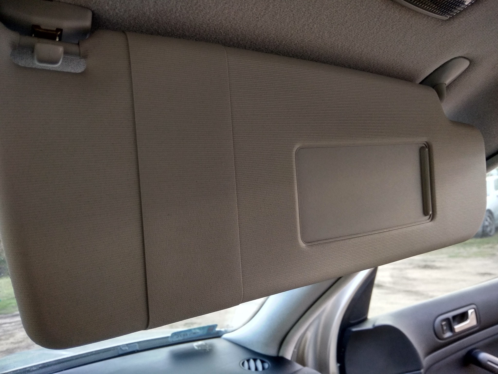 Szary daszek przeciwsłoneczny w samochodzie z widocznym uchwytem mocującym i lusterkiem, wnętrze pojazdu z elementami deski rozdzielczej i drzwi.