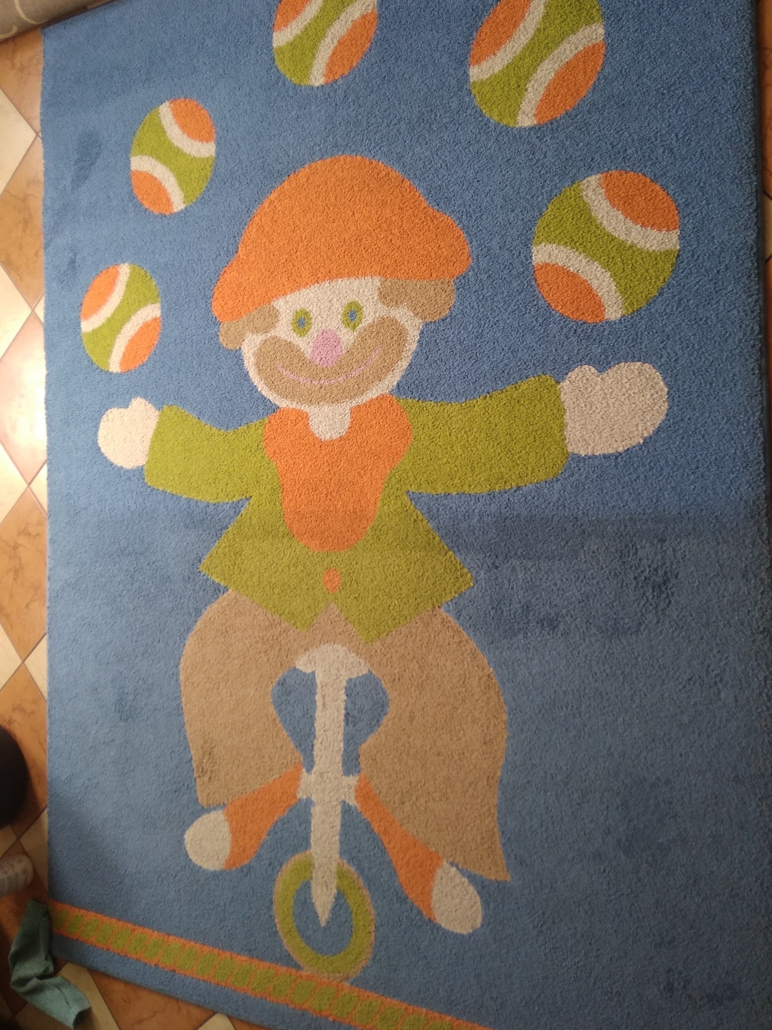 Niebieski dywan z wzorem klauna żonglującego piłkami na monocyklu, widoczne plamy i zabrudzenia.