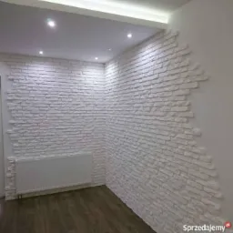 Ściana wykończona dekoracyjną białą cegłą w Olesno, z widocznym grzejnikiem i fragmentem drewnianej podłogi, podświetlona listwą LED w suficie.