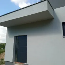 Fragment nowoczesnego domu z szarą elewacją, widoczna boczna ściana z drzwiami wejściowymi, zadaszenie nad wejściem i fragment okna. Elewacja o delikatnej strukturze, w tle fragment nieba i zieleni.