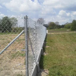 Długie, ocynkowane ogrodzenie z siatki leśnej, wsparte na słupkach z rur, rozciąga się wzdłuż granicy działki. Słoneczny dzień, widok na łąkę i drzewa w tle.