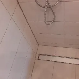 Fragment łazienki z beżowymi płytkami, widoczny odpływ liniowy i wąż prysznicowy zwisający z góry.