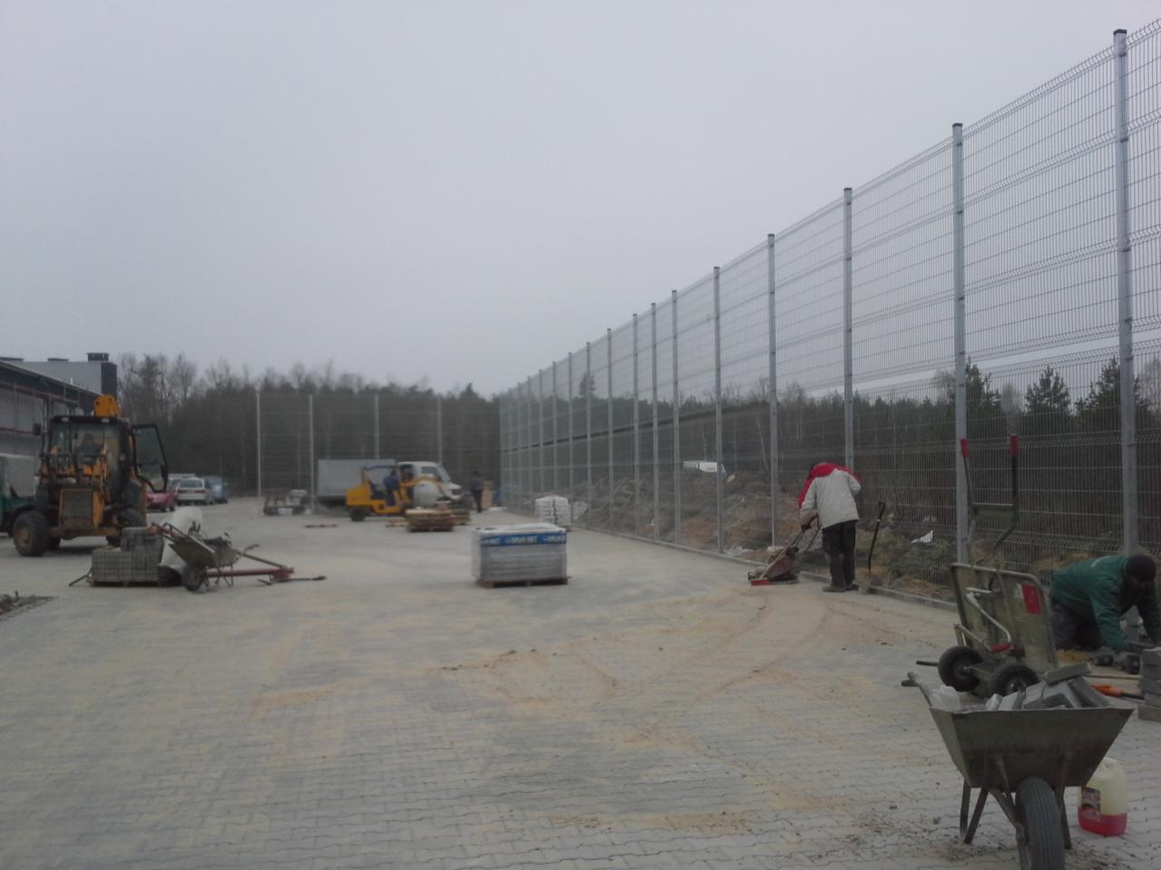 Montaż panelowego ogrodzenia na posesji, widoczne narzędzia i materiały budowlane, w tle las i pochmurne niebo. Prace w toku.