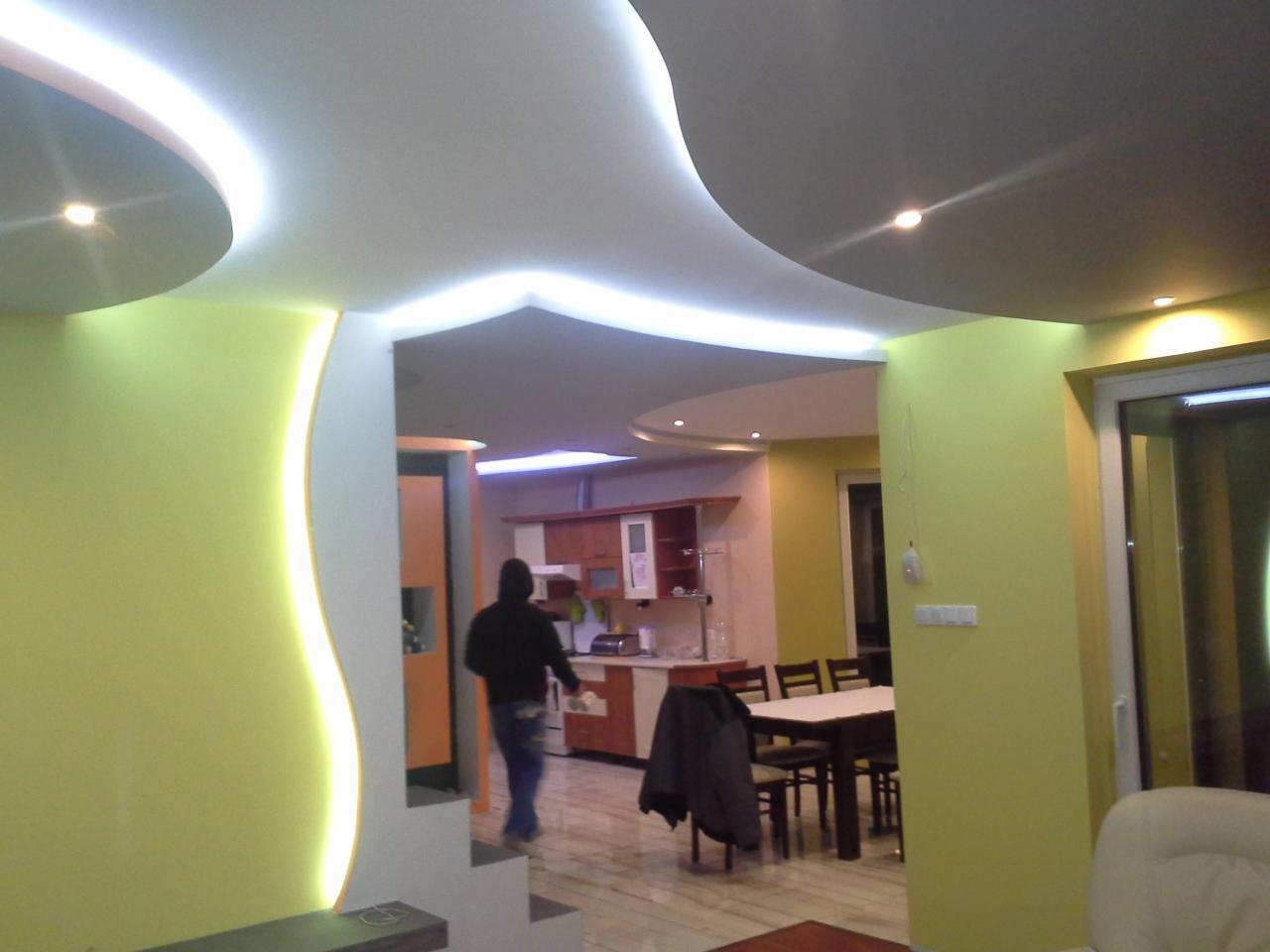 Nowoczesne wnętrze z podwieszanym, podświetlanym sufitem w kształcie fali. Żółte ściany i widoczna kuchnia w tle. Osoba w oddali przechodzi do innego pomieszczenia.