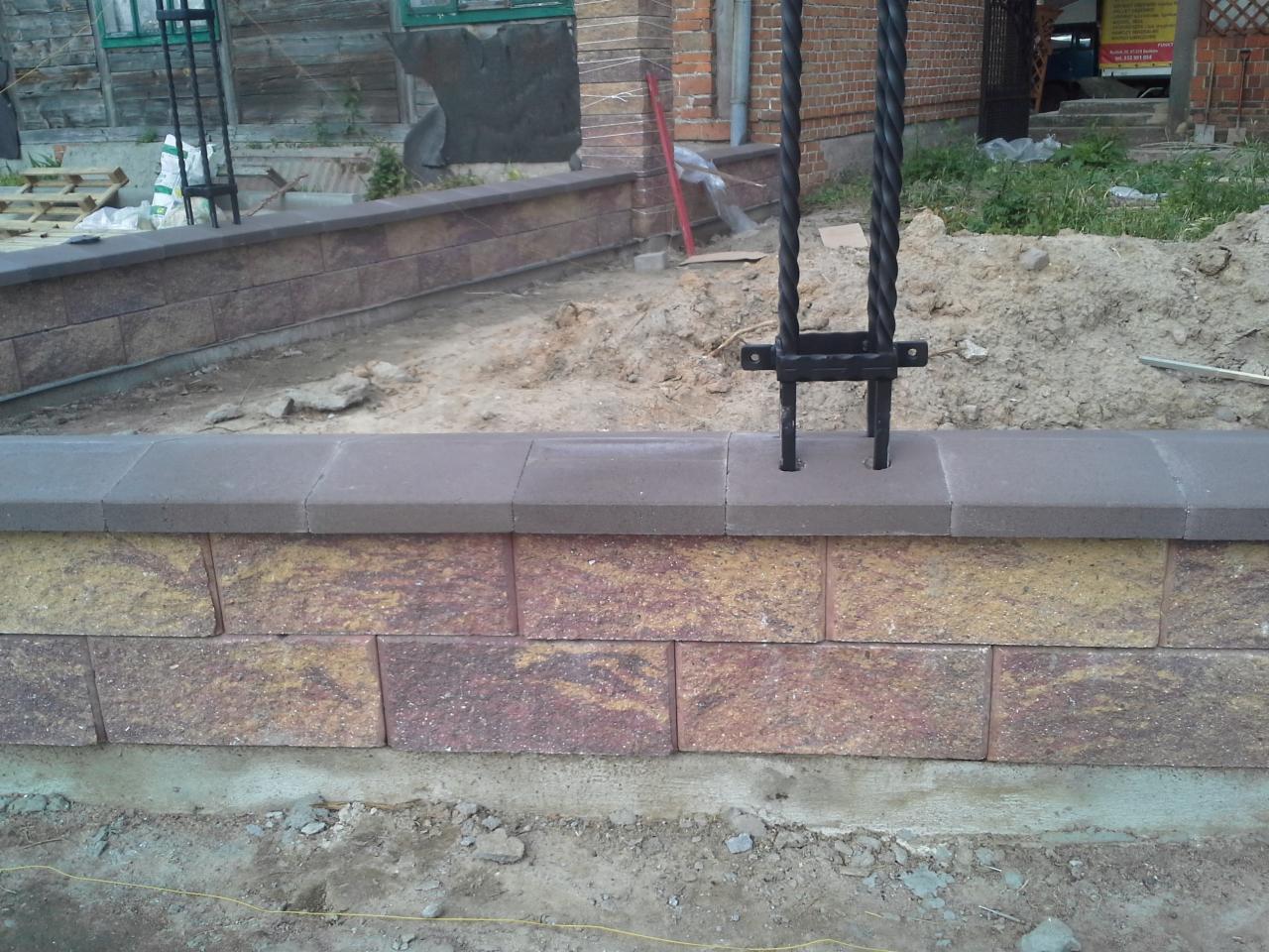 Fragment murku z bloków betonowych w odcieniach beżu i brązu, z wmontowanymi elementami metalowymi, przygotowany do montażu. Widoczne prace ziemne w tle.