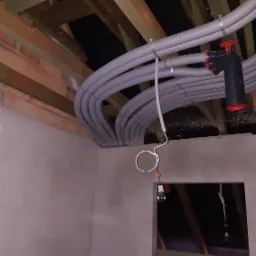 Wiązka szarych rur karbowanych prowadzona pod drewnianą konstrukcją stropu, zabezpieczona metalowymi obejmami, z widocznym czarnym elementem z czerwonymi akcentami, surowa ściana i tymczasowe...