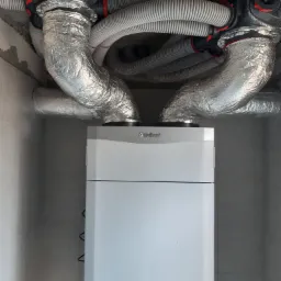 Biała jednostka rekuperacji Vaillant zamontowana w pomieszczeniu, z widocznymi izolowanymi kanałami wentylacyjnymi pod sufitem.