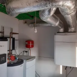Zainstalowane urządzenia grzewcze Vaillant w pomieszczeniu: pompa ciepła, zbiornik buforowy, naczynie przeponowe oraz izolowane rury wentylacyjne pod sufitem.