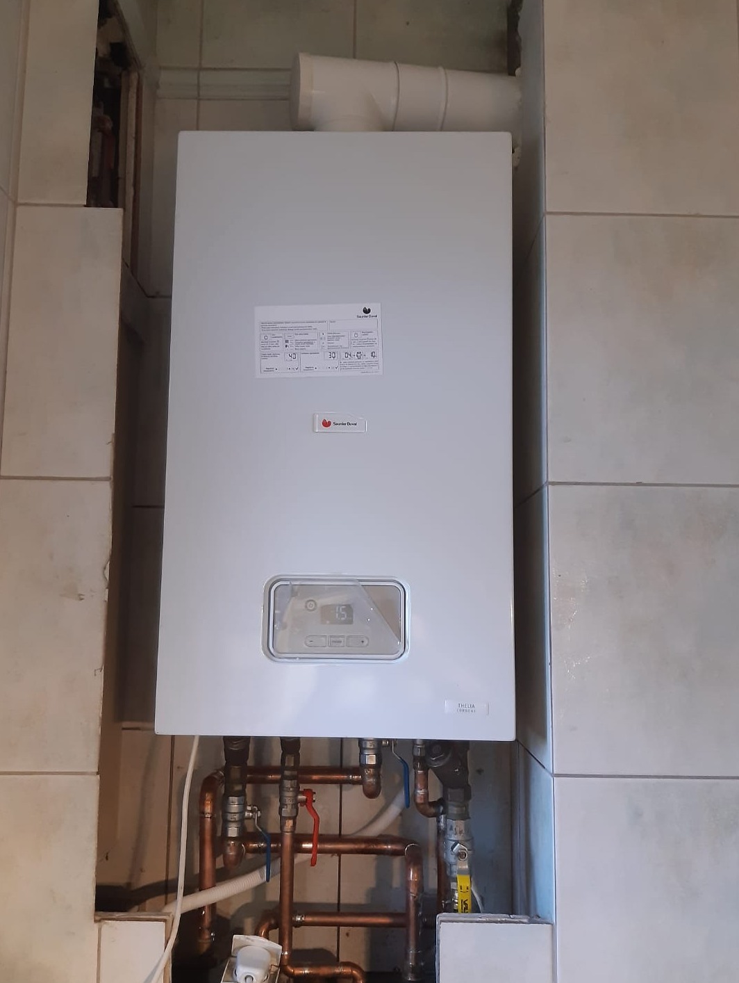 Biały piec gazowy marki Saunier Duval zamontowany na ścianie z beżowych płytek, widoczne miedziane rury z zaworami i biały przewód odprowadzający spaliny.