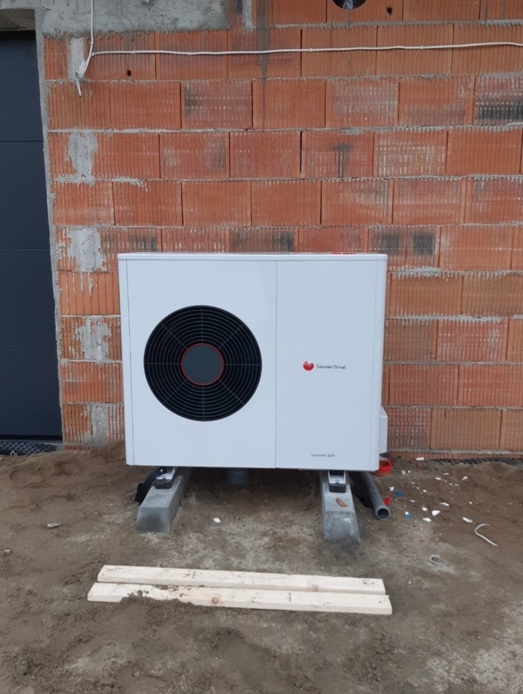 Biała pompa ciepła Saunier Duval ustawiona na betonowych bloczkach przed surową ścianą z czerwonej cegły dziurawki, widoczne przewody i elementy instalacji.