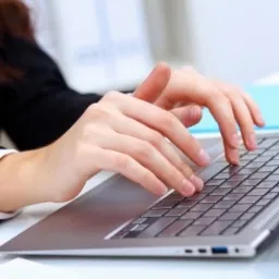 Dłonie kobiety w czarnej marynarce piszą na klawiaturze laptopa; widok z bliska na palce i klawisze.