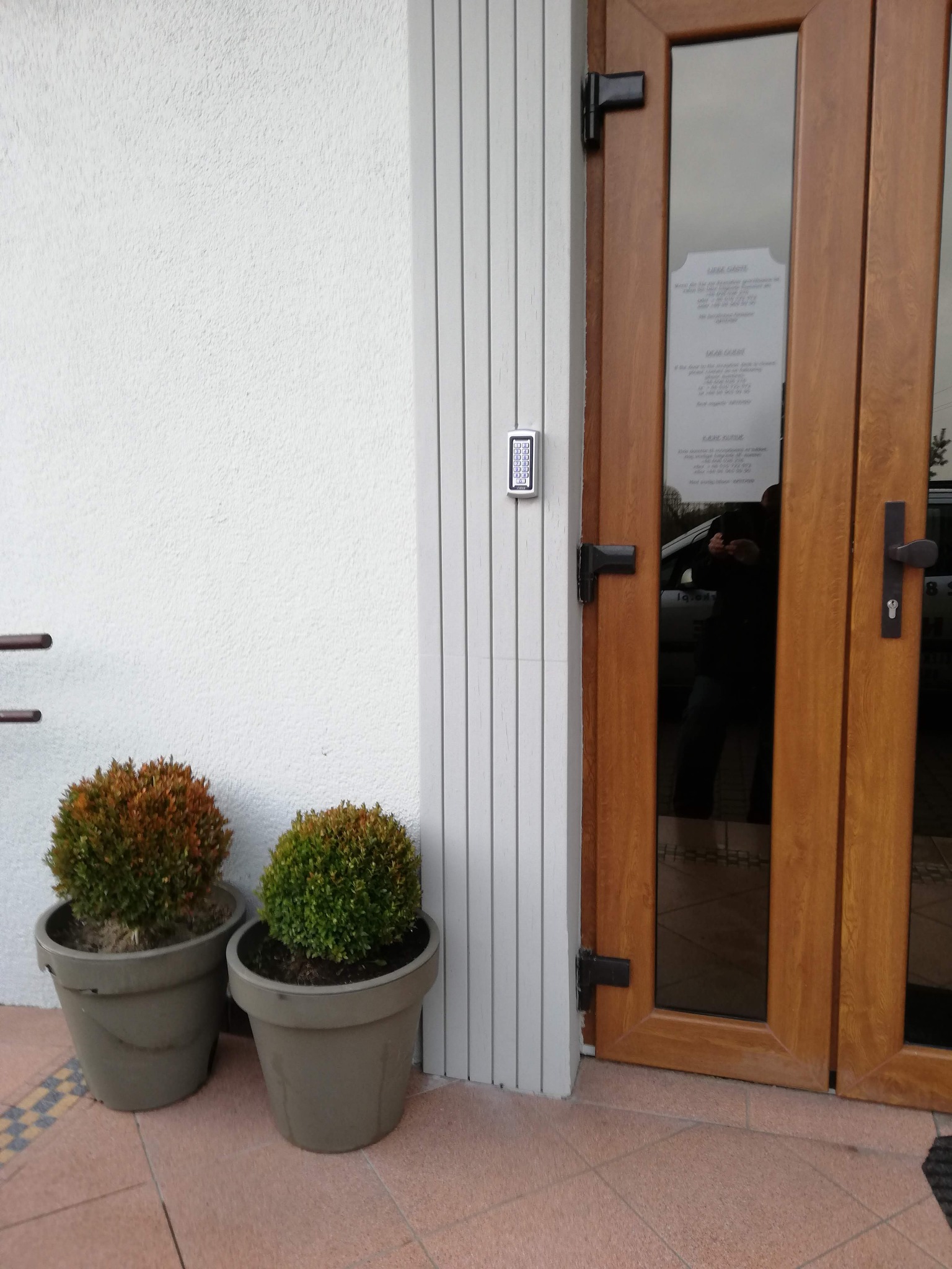 Zewnętrzny panel domofonowy z klawiaturą numeryczną zamontowany obok drewnianych drzwi wejściowych z szybami, dwie okrągłe rośliny w donicach na pierwszym planie.