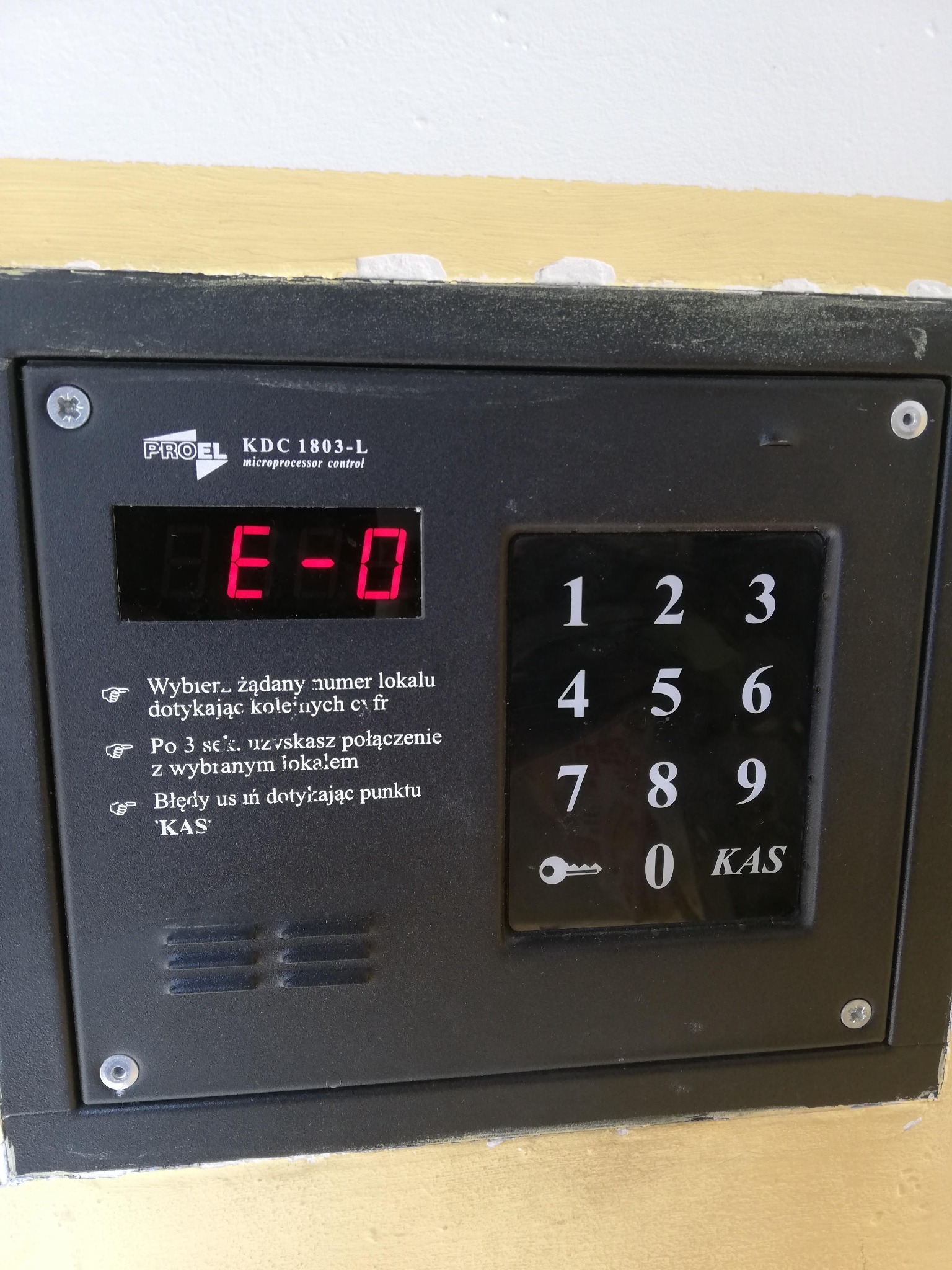 Panel domofonu PROEL KDC 1803-L z podświetlanym wyświetlaczem pokazującym 'E-0' i klawiaturą numeryczną, zamontowany na pomalowanej ścianie.