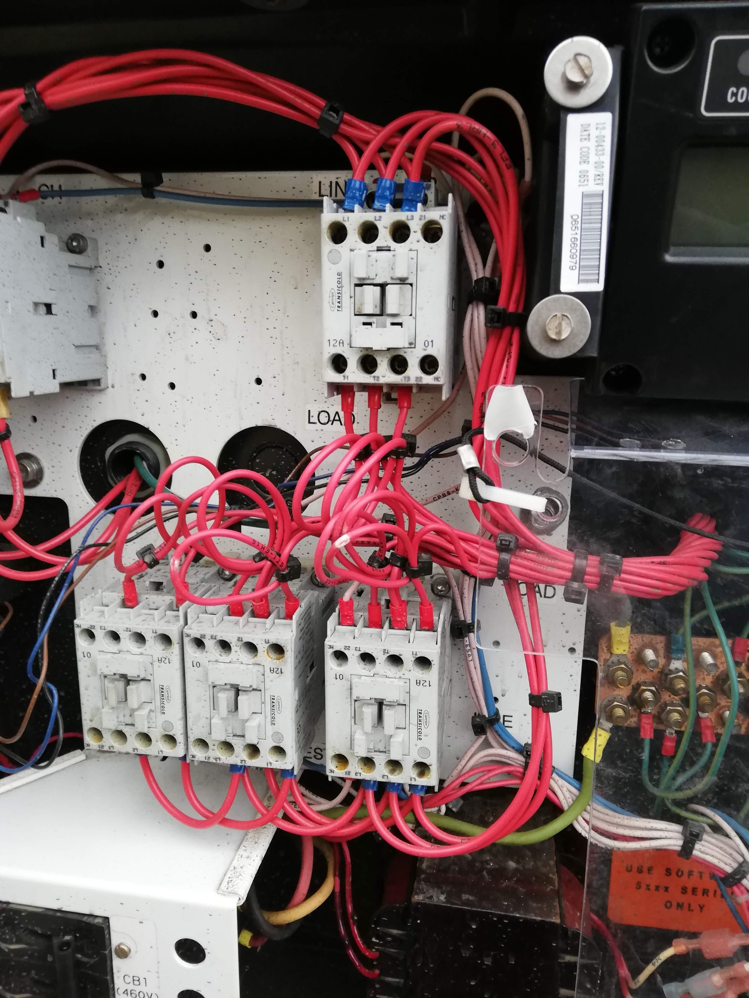 Rozdzielnica elektryczna z widocznymi stycznikami, zabezpieczeniami i przewodami w kolorze czerwonym, zamocowanymi opaskami zaciskowymi, z elementami opisanymi kodami i oznaczeniami.