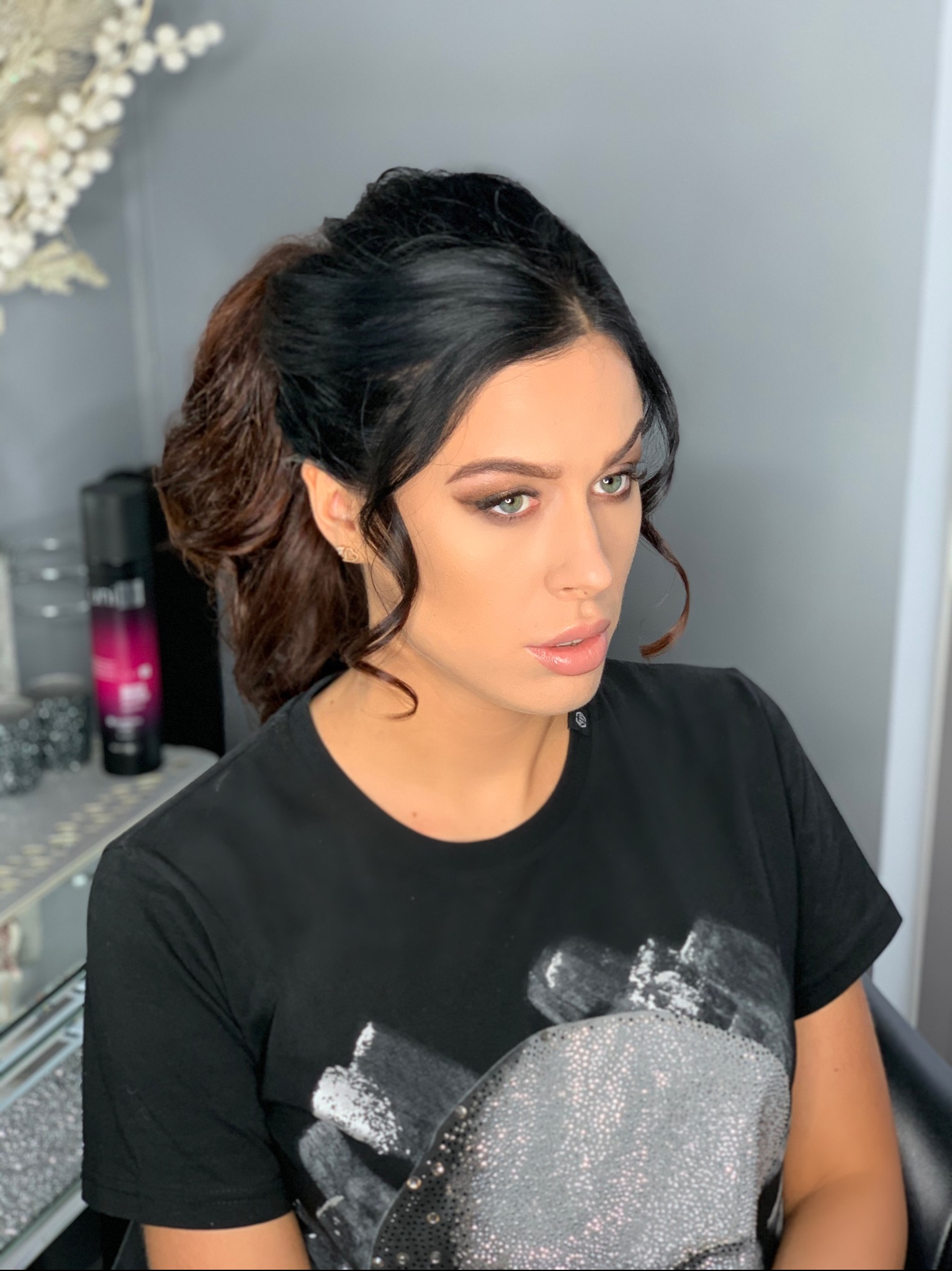 Portret kobiety z brązowo-czarnymi włosami upiętymi w koński ogon, delikatny makijaż oczu w odcieniach brązu, błyszczące usta i kolczyk w kształcie logo Chanel.