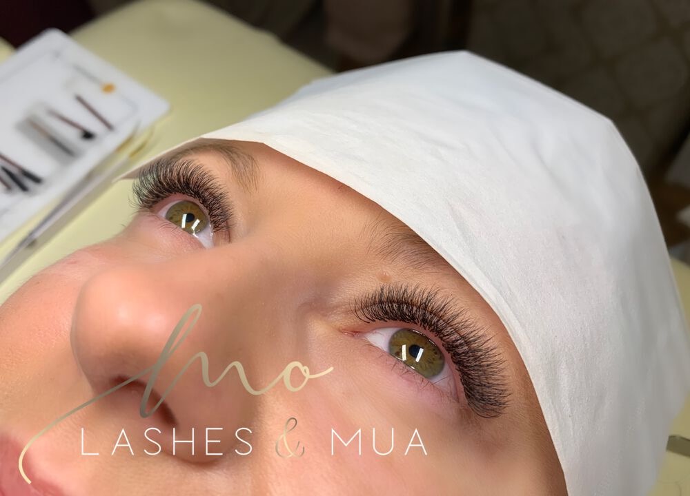 Zbliżenie na oko z wyraźnie przedłużonymi, gęstymi rzęsami w stylu 2D/3D; widoczne złote refleksy w tęczówce, biała chusta na głowie i logo firmy w dolnej części zdjęcia.