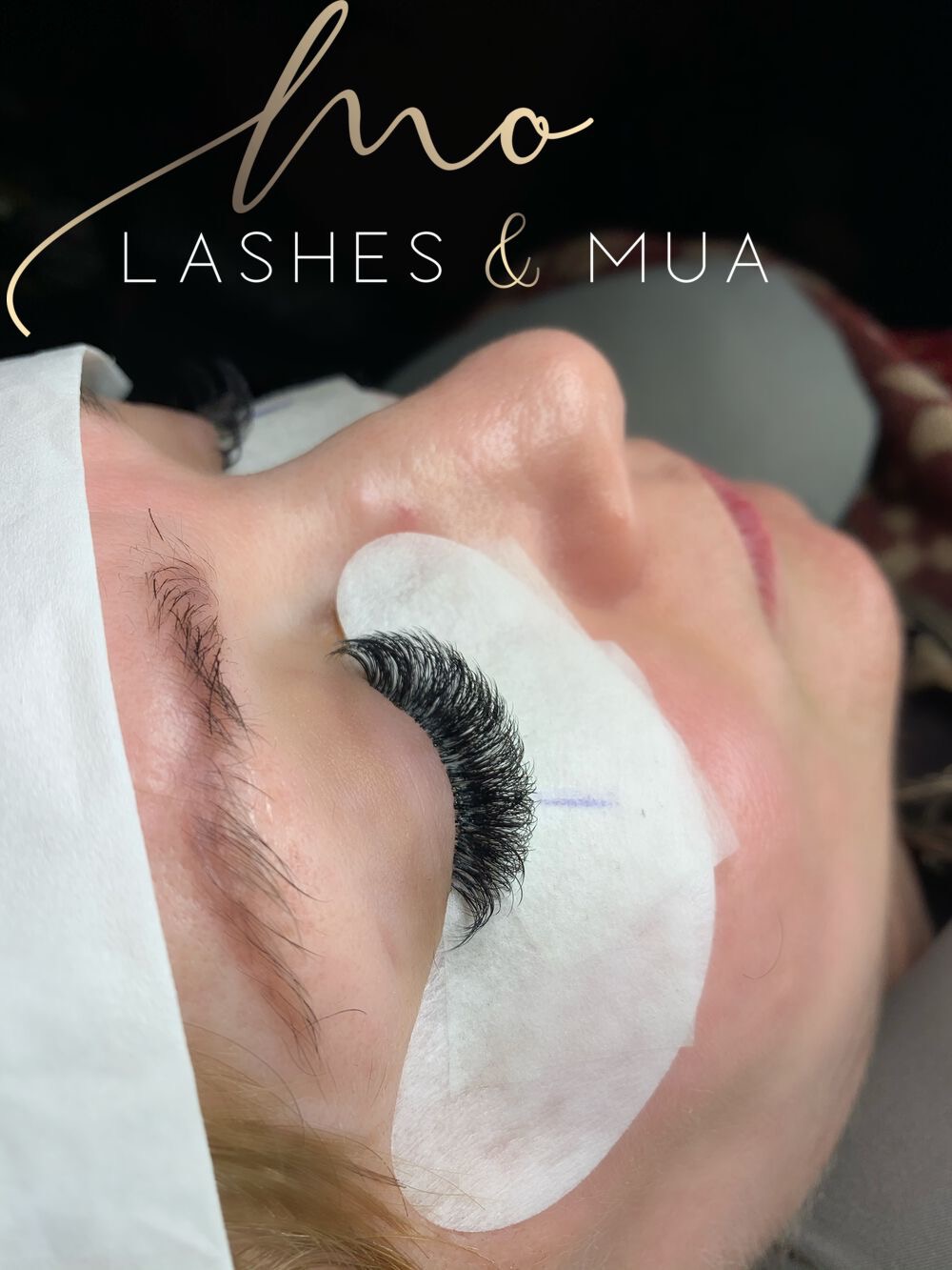 Zbliżenie na oko kobiety z założonymi, gęstymi sztucznymi rzęsami; widoczna ochronna podkładka pod okiem i logo 'ho LASHES & MUA' w górnej części kadru.