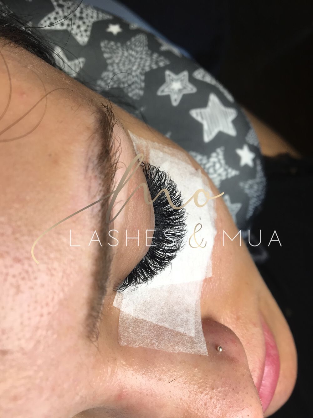 Zbliżenie na oko z przedłużonymi, czarnymi rzęsami, zabezpieczonymi taśmą kosmetyczną, z widocznym piercingiem w nosie i logo 'Lashess & MUA' w rogu.