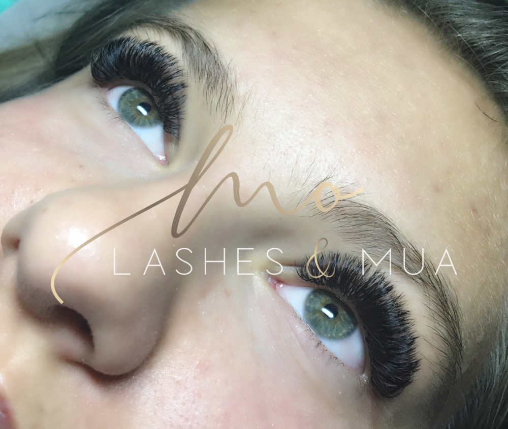 Zbliżenie na oko z gęstymi, czarnymi, przedłużonymi rzęsami w stylu 'Russian Volume' i zieloną tęczówką, widoczne logo 'LASHE & MUA' na twarzy.