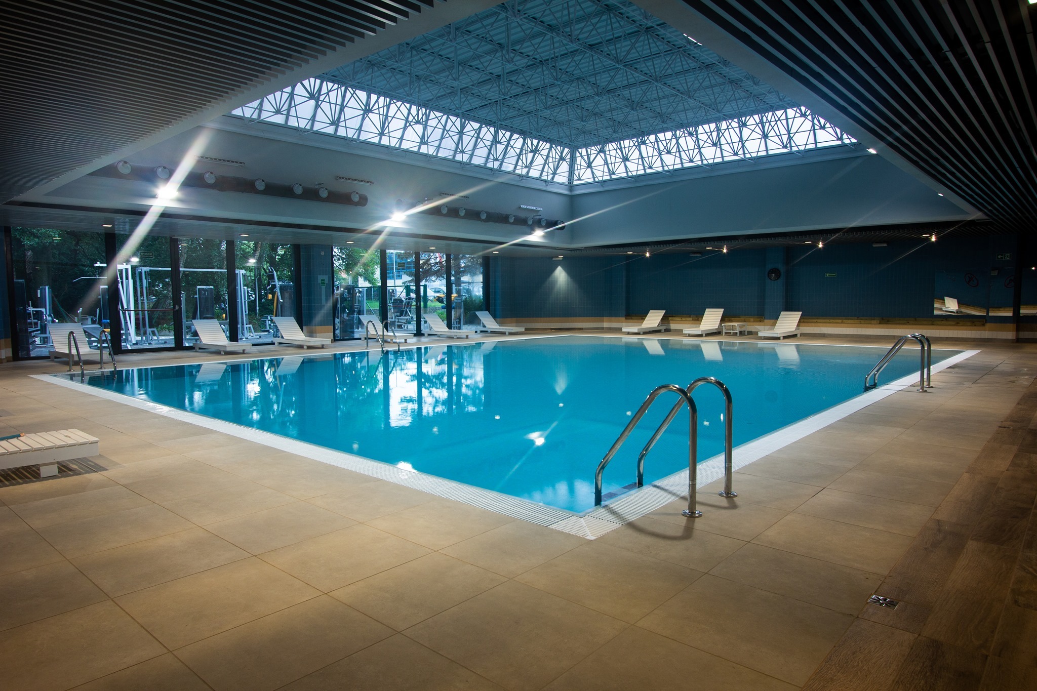 Kryty basen z turkusową wodą, otoczony leżakami i siłownią, oświetlony naturalnym światłem przez przeszklony sufit.