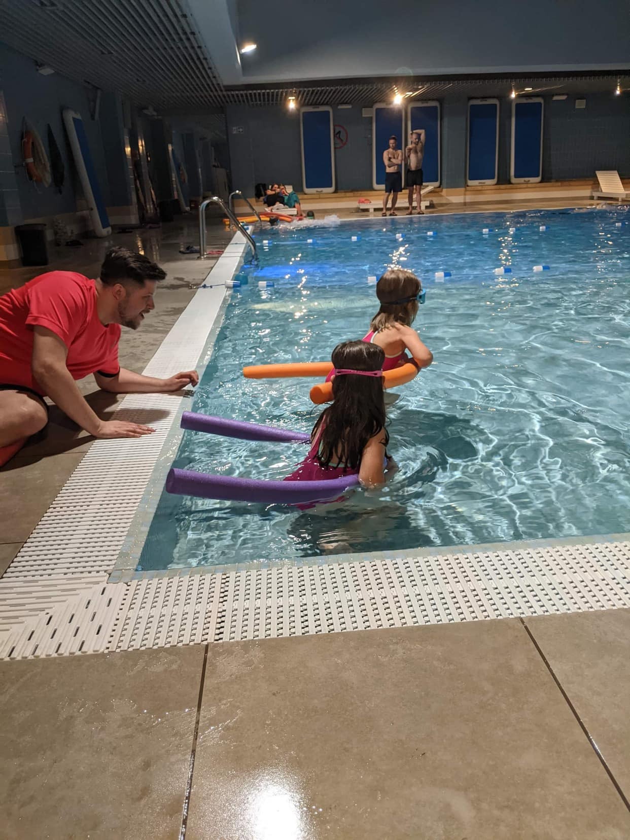 Instruktor pływania w czerwonej koszulce obserwuje dwie dziewczynki w różowych strojach kąpielowych, używające piankowych makaronów do pływania w basenie krytym.